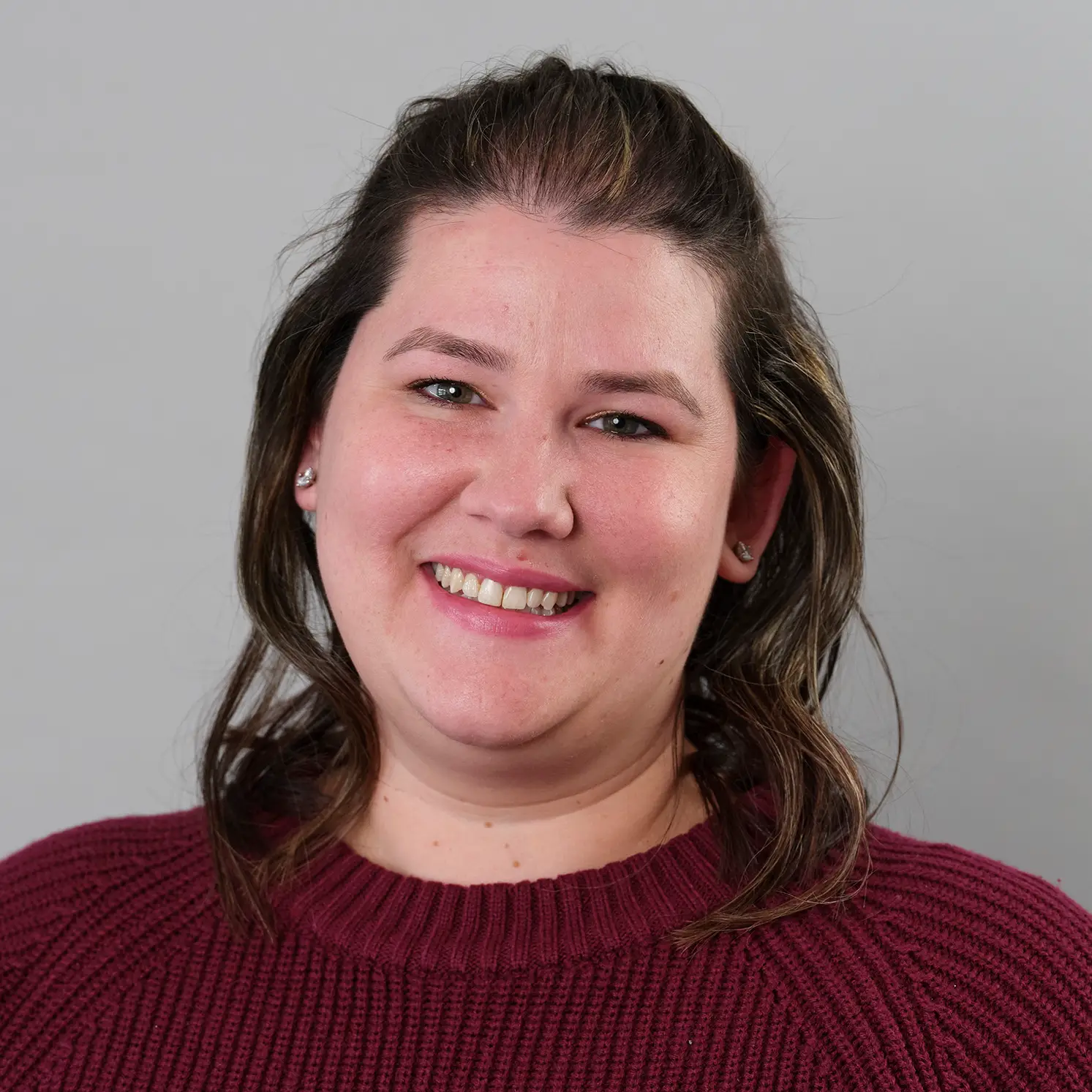 A woman with shoulder-length brown hair smiles at the camera. She is wearing a maroon sweater and has a neutral gray background behind her.