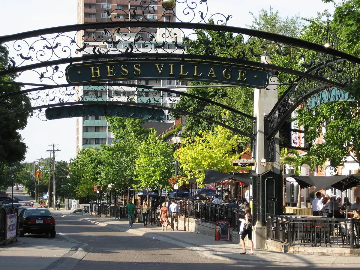 A street scene at Hess Village, with an ornate wrought iron archway displaying the name "Hess Village". People are walking along the sidewalk, and there are outdoor seating areas from nearby restaurants. Institutional investors admire the flourishing locale as trees and buildings line the street on a sunny day.