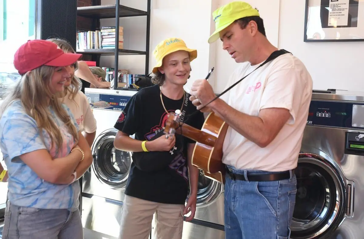 Three people, two wearing yellow and red caps, watch as a man, also wearing a yellow cap, signs a ukulele at the Hamilton Community laundromat. They are standing in front of washing machines.