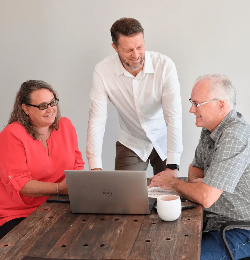 Three people at a wooden table: a woman in a red top sits on the left, a man in a white shirt stands in the middle, and a man in a gray shirt sits on the right. They are engaging in a discussion, with a laptop and a cup on the table.