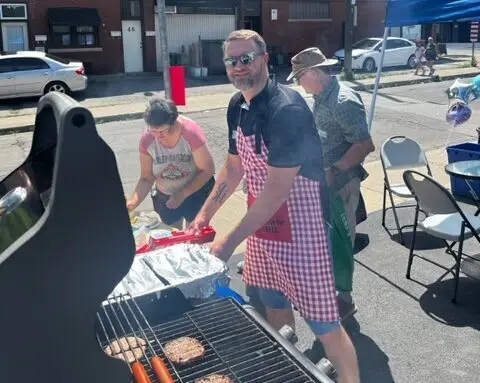 A man in sunglasses and a red and white checkered apron cooks burgers and hotdogs on a grill at an outdoor Hamilton Community event. A woman nearby is preparing food on a table, and an older man stands behind her. They are on a sunny street with cars and buildings in the background.